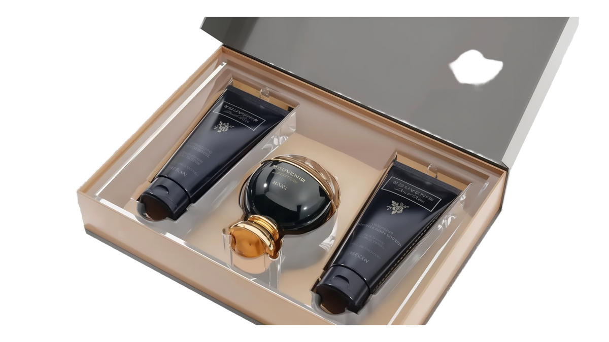 The image shows an elegant gift set by Afnan Perfumes from their "SOUVENIR Desert Rose" collection. The set is displayed in a beige presentation box with a clear lid, revealing its contents. Inside, there is a dark, glossy perfume bottle with a spherical design and a golden cap, positioned centrally. On either side of the perfume bottle are two matching dark tubes. Both tubes and the perfume bottle feature the "SOUVENIR Desert Rose" branding in elegant gold lettering