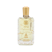 The image displays an elegant, clear glass perfume bottle adorned with artistic golden lines and splashes against a soft, off-white backdrop. The bottle is labeled "3SHQ INTENSE" in a refined font, with the "DKHAN FRAGRANCES" emblem prominently featured beneath it. A gold-colored cap sits atop the bottle, complementing the golden accents. The fragrance liquid has a light, amber tint, suggesting a sense of warmth and richness.