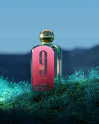 The image shows a rectangular glass perfume bottle with a pink front label and a gold spherical cap, placed on a surface with greenery. The label features a large black "9" and "am" in black font, with "pour femme" in smaller script below it. The text "AFNAN" is visible at the bottom of the label.  The bottle is set against a moody background with a blue gradient, evoking a night-time scene, and is illuminated from above, creating a soft reflection on the cap.