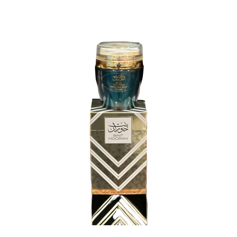 The image shows a product named "Bint Hooran Hand and Body Lotion." The lotion comes in a dark teal container with a clear lid and is placed on top of a matching box adorned with elegant geometric patterns and Arabic calligraphy. The box prominently features the product name "Bint Hooran" along with intricate designs in silver and gold. The luxurious and sophisticated packaging suggests a high-quality skincare product.
