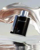 The image displays a sleek and contemporary perfume bottle floating against an artistic backdrop. The bottle has a shiny black glass exterior with a smooth, reflective silver cap. In the background, there is a fluid and dynamic metallic structure with a rose gold finish that swirls around the bottle, creating a sense of movement. Below the bottle, there appears to be a reflective surface, reminiscent of water, adding to the overall ethereal and high-end feel of the presentation. 