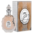 The image features a perfume bottle and its packaging:  The perfume bottle is crafted from multi-faceted glass with a pale pink tint and a decorative surface texture. It is adorned with a clear glass stopper that has a detailed pattern and a metallic chain with a charm attached to the neck. The label on the bottle has Arabic calligraphy and the name "ROUAT AL MUSK" in English.