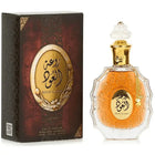 The image shows a perfume bottle and its box:  The perfume bottle is made of clear glass with an ornate design and features an amber-colored central area with decorative elements. Arabic calligraphy is displayed in the central area, with "ROUAT AL OUD" inscribed in English below it. The bottle has a clear, intricately designed stopper and a golden chain with a charm around its neck.