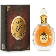 The image shows a perfume bottle and its box:  The perfume bottle is made of clear glass with an ornate design and features an amber-colored central area with decorative elements. Arabic calligraphy is displayed in the central area, with "ROUAT AL OUD" inscribed in English below it. The bottle has a clear, intricately designed stopper and a golden chain with a charm around its neck.