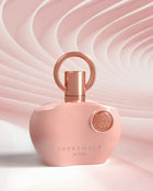 The image features a pink perfume bottle with a glossy finish, set against a textured background with a wavy, fabric-like pink design that creates a sense of motion. The bottle has a unique shape with rounded edges and a flat profile. It is adorned with a rose gold-colored ring-shaped cap and matching spray nozzle. The lighting highlights the bottle's curves and the emblem's detail. 