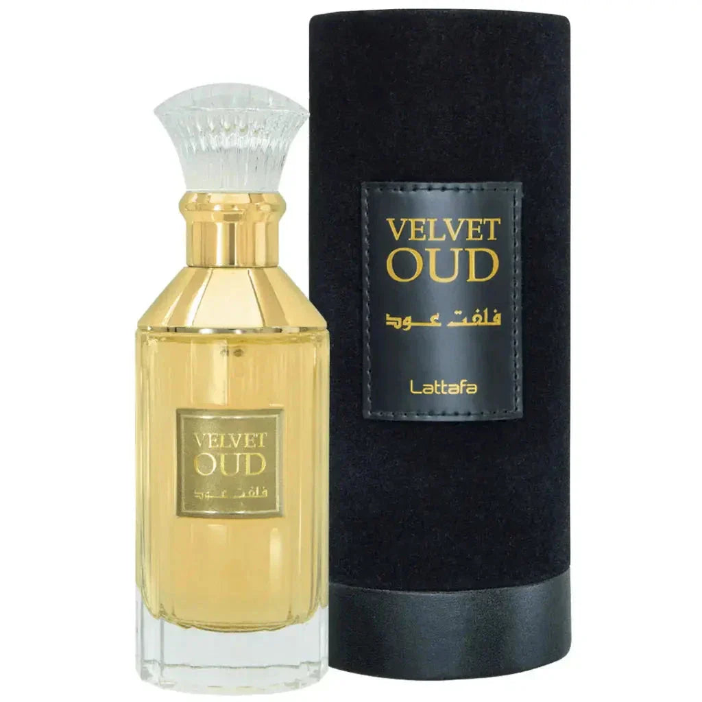The image features a perfume bottle alongside its cylindrical container:  The perfume bottle is rectangular, crafted from clear glass with a gold-colored liquid visible inside, indicative of the perfume it contains. A white, bristle-like applicator tops the bottle, resembling a traditional shaving brush, and there's a clear, rounded cap above it. The label on the bottle is gold with the name "VELVET OUD" in bold, along with Arabic script.